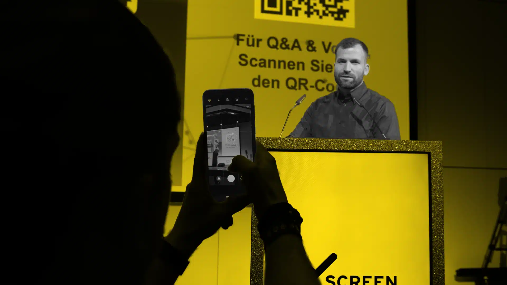 Eine Besucherin scannt einen QR-Code, welcher auf einer Leinwand hinter der Bühne und auf der Bühne im Rednerpult angezeigt wird