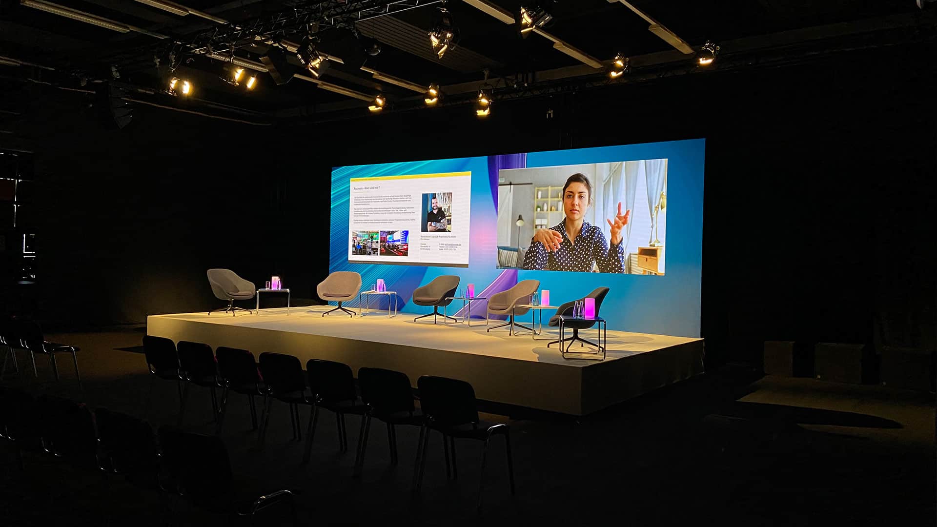 Bühne mit 5 Sesseln vor einer LED Wand mit einem Bildausschnitt einer Kamera und eines weiteren Ausschnittes einer Powerpoint Präsentation