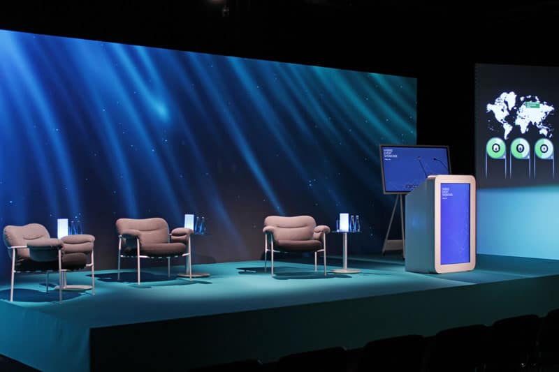 eine Bühne mit einer LED Wand und davor ein Rednerpult, Sessel und ein Flipchart